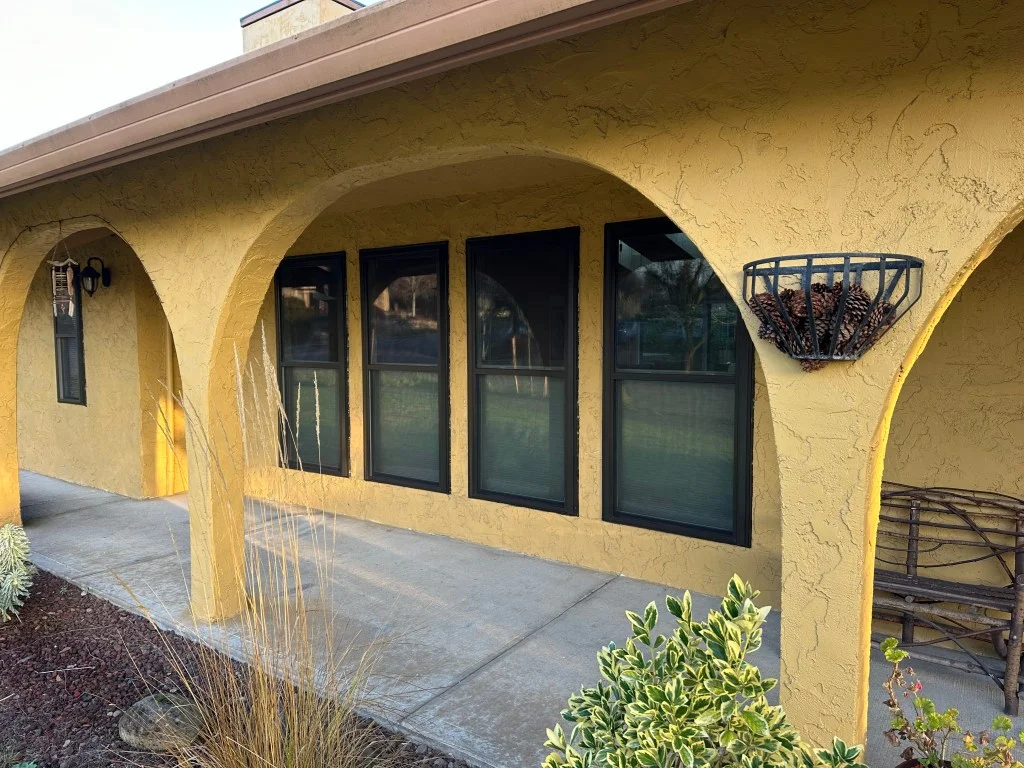 Four tall dark-framed windows set in yellow stucco wall under arched covered walkway with concrete floor