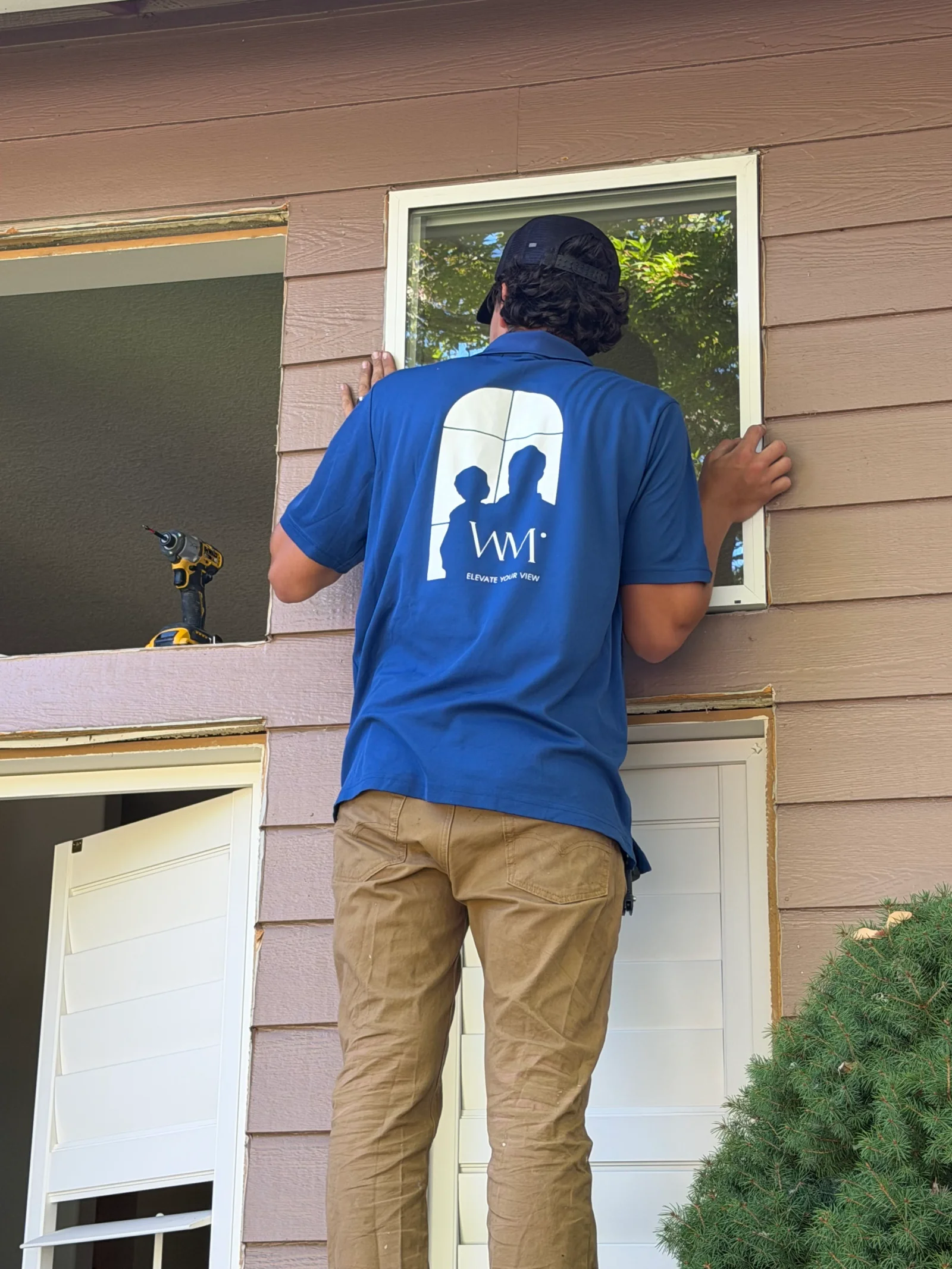 Technician installing white-framed window into tan lap siding wall with cordless drill on sill